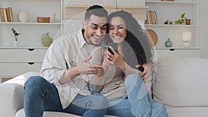 Overjoyed young hispanic married couple looking at smartphone screen sitting on couch getting message with good news