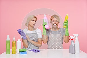 Overjoyed young pretty blonde housewife keeping household chemicals in raised hands and looking cheerfully at camera, sitting over