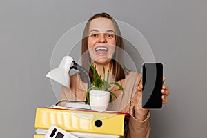 Overjoyed young pretty woman wearing beige jacket holding documents and her office stuff, showing mobile phone with empty display