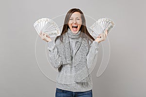 Overjoyed young woman in gray sweater scarf screaming, holding lots bunch of dollars banknotes, cash money isolated on