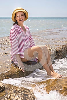 Overjoyed young woman laughing sitting on stone beach surrounded by water splashes foamy waves