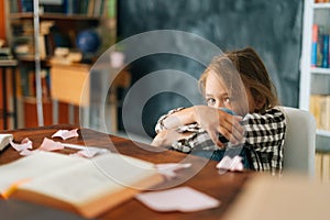 Overload upset primary child school girl sitting alone hugging knees in front of desk with difficult homework looking at