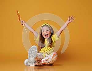 Overly excited kid girl in yellow t-shirt and colorful leggings sits on floor with big lollipop and holding hands up