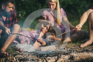 Overnight camping in summer. Cooking sausages for dinner in camp. Blurred two couples sitting by campfire