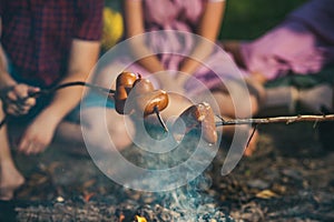 Overnight camping in summer. Cooking sausages on sticks over flames of campfire. Two couples sitting by bonfire