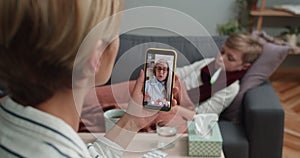 Overshoulder view of mother holding mobilephone and communicating with mature doctor. Mother having online medical