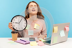 Overtime work. Fatigued upset woman employee sitting at workplace office, all covered with sticky notes and holding big clock