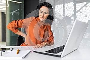 Overtired hispanic woman working late, business woman having severe back pain, female worker inside office sitting at photo