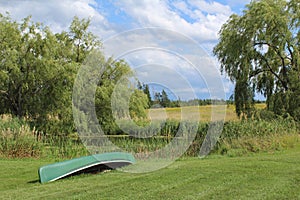 An overturned canoe lying by a pond with willow trees and a grassy field in the background on a sunny summer day