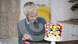 Overview footage of woman with short grey hair decorates small white cake with colorful flowers on white modern kitchen