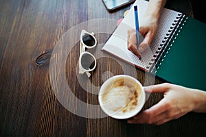 Overview of woman`s hands writing in a notepad, holding coffee.