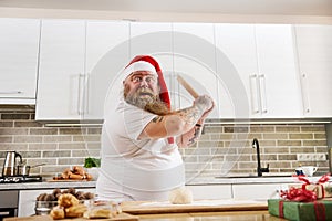 An overweigh man in Santa Claus hat with tattooed arms wearing a white T-Shirt, holding a wooden rolling pin on hands like a