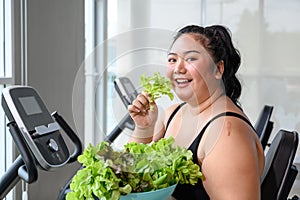 Overweight Asian female happy to eat organic green vegetable salad for healthy in gym. Weight loss workout, healthy lifestyle