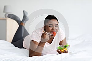 Overweight black woman eating tasty vegetable salad, lying on bed at home. Healthy nutrition for weight loss
