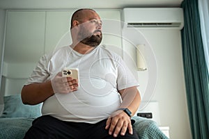 Overweight body positive bearded man with phone and fitness tracker thinking on fatness problem
