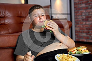 Overweight boy with fast food on floor near sofa at home, eating junk food