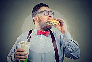 Overweight business man eating with appetite a burger holding a can of soda drink