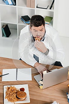 overweight businessman eating junk food while working with laptop