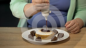 Overweight female pouring lots of chocolate dressing over pastry, obesity issue