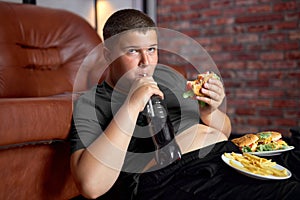 Overweight kid with fast food on floor near sofa at home, eating junk food