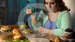 Overweight lady exercising and sadly looking at junk food, obesity problem