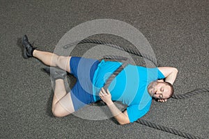 Overweight man is lying on the floor exhausted after performing battle rope exercise in the fitness gym