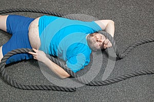 Overweight man is lying on the floor exhausted after performing battle rope exercise in the fitness gym