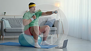 Overweight man sitting on fit ball and exercising with dumbbells at home, watching fitness lesson on laptop