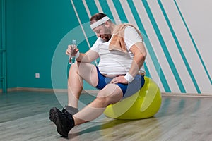 Overweight man is sitting on a fitness ball exhausted after a hard training in group fitness classes. Fat man is