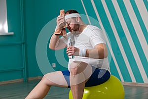 Overweight man is sitting on a fitness ball exhausted after a hard training in group fitness classes. Fat man uses a