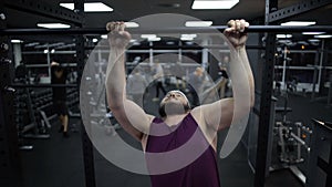 Overweight man trying to pull up on sport bar, weak body muscles, gym training