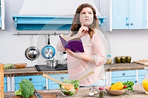 overweight pensive woman with cookery book standing in kitchen photo