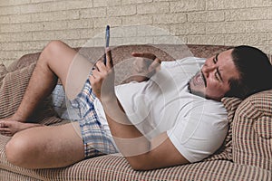 A overweight and unshaven man laughs at some funny videos streaming on the cellphone while lounging on the sofa during a lazy day