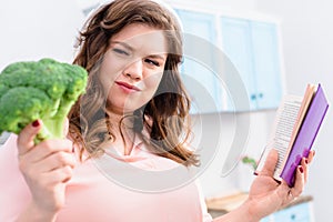 overweight woman in headphones with cookery book looking at fresh broccoli in hand photo