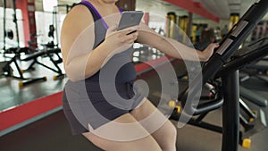 Overweight woman riding slowly stationary bike and chatting on mobile phone