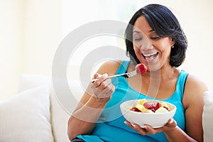 Overweight Woman Sitting On Sofa Eating Bowl Of Fresh Fruit