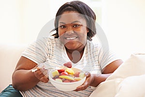 Overweight Woman Sitting On Sofa Eating Bowl Of Fresh Fruit