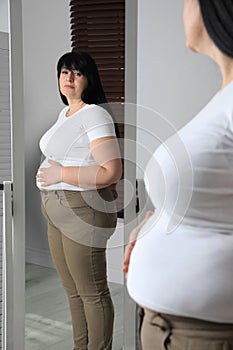 Overweight woman in tight t-shirt and trousers near mirror at home