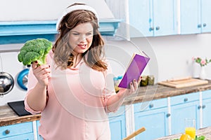 overweight young woman in headphones with fresh broccoli and cookery book photo