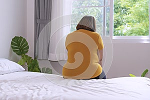 Overweight young woman sitting on white bed at home. Upset female suffering from extra weight. Obesity unhealthily concept