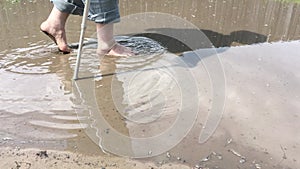 Overweighted senior man with walking stick walking barefoot through pool on an earth road at summer season