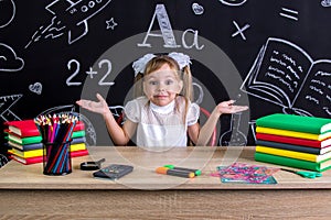 Overwhelmed smiling schoolgirl sitting at the desk with books, school supplies, holding her both arms with palms upto