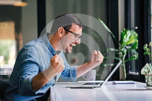 Overwhelmed by success young cheerful adult casual businessman or entrepreneur smiling and vividly celebrating with raised hands