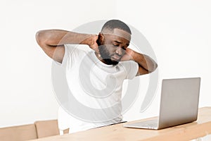 Overworked African American employee suffering from neck pain sits with laptop in home office and massages his own neck.