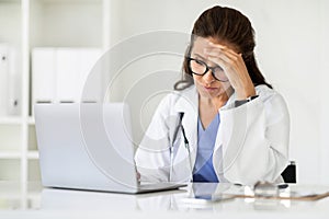 Overworked tired mature woman doctor working on laptop at clinic