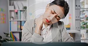 Overworked tired young Asia businesswoman sit on desk with laptop massage stiff neck to relieve pain in muscles at office. Female