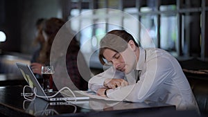 Overworking Caucasian man falling asleep on bar counter with blurred interracial couple meeting at background