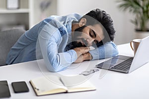 Overworking Concept. Tired Young Indian Man Napping At Desk With Laptop