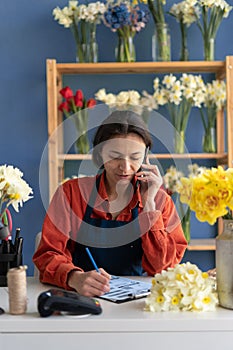 Own business concept. Happy businesswoman talking on mobile phone in small floral shop and writing order details