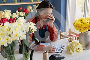 Own business concept. Happy businesswoman talking on mobile phone in small floral shop and writing order details